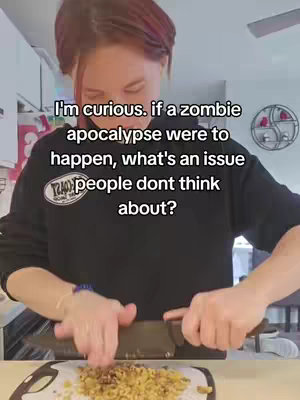 In a kitchen setting, a person with red-tinted hair is shown preparing food while text overlays appear on the screen. The person is dressed in a black sweatshirt and is focused on cutting something with a knife. The general layout includes the person as the foreground while the kitchen acts as the background.