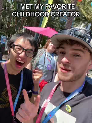 A selfie-style image featuring three people. The main subject is a man wearing a hat and smiling, with sunglasses perched on his hat. To his left, another man with glasses is grinning widely, pointing towards the camera. A third person with curly hair is visible in the background. The text overlay reads 'I MET MY FAVORITE CHILDHOOD CREATOR.' The overall composition is bright and appears to be shot outdoors, likely in a public setting.