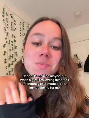 A selfie-style video with a woman speaking directly to the camera. The background is a simple, slightly cluttered room. The woman has long, wavy brown hair and is wearing dark clothing. Text overlay is on the lower part of the screen.