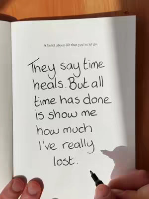 An open book on a wooden surface, displaying handwritten text. The text is in black ink, with the writer's hand holding the pen in the lower right corner of the image. The book's pages are cream-colored and the lighting appears soft and natural.