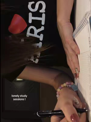 The cover image shows a close-up shot with a person holding a pen over a notebook. The person is wearing a black shirt with an 'I ❤️ PARIS' logo. On the right edge of the frame, a hand with long, manicured nails and a bracelet is visible. The image is slightly rotated in a vertical orientation, with text overlayed at the bottom left that says 'lonely study sessions !'.