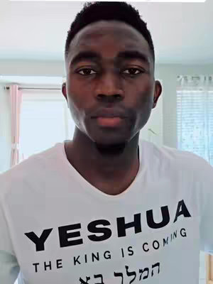 A medium close-up shot of a young Black man looking directly at the camera. He is wearing a white t-shirt with bold black text that reads 'YESHUA' in large capital letters, and below, in a smaller font, 'THE KING IS COMING' and Hebrew characters. His face has a serious expression, with short, neatly styled black hair. The background shows a soft-focus interior space with light-colored walls, a window with sheer curtains, and the top of a closed door.