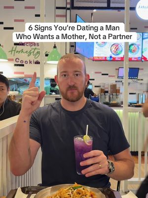 A man with fair skin and red hair, wearing a dark blue t-shirt, is seated at a table indoors. He is holding a purple drink with a straw and lime slice. He is pointing his right index finger upward. A bowl of food and a stainless steel tray are in the foreground, while blurred restaurant scenes in the background include advertisements.