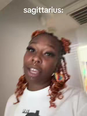 A selfie-style video featuring a woman with orange and brown curly hair styled in space buns, a nose ring, and heavy eye makeup. The background is a simple light-colored wall with a glimpse of what looks like a corner.