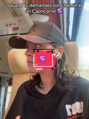 A selfie-style view of a person in a train. The person, on the left side of the frame, is wearing a black t-shirt, a cap, and earbuds. A colorful graphic with a zodiac symbol is on the face. The background shows the interior of a train car with seats.