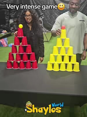 The image shows two people, a woman and a man, standing behind a black table. Both people are holding a structure made of plastic cups. The woman is on the left side, building a tower of red cups with a yellow ball on top. The man is on the right side, holding a similar structure made of yellow cups with a red ball on top. Above, the text "Very intense game" is displayed with a smiling emoji. The background is a blurry indoor setting, and the logo of "Shaylos World" is at the bottom.