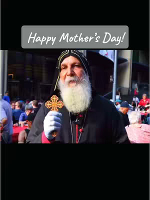 The image is a vertical shot featuring a man with a white beard, wearing a dark robe and a black hood. He is holding a gold cross and is positioned in front of a crowd. The top of the image has a rectangular banner with the text "Happy Mother's Day!" in white. Background includes buildings and people.