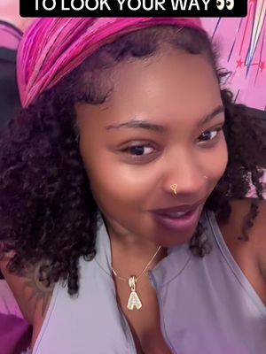 A selfie-style shot featuring a woman smiling, gazing slightly off to the side. She has curly dark hair, a pink headscarf, and is wearing a gray top. She has a small nose ring and a necklace with a pendant. The background is blurred, with pink and white colors faintly visible.