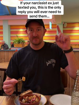A man in a black t-shirt and a baseball cap is seated at a table, likely in a restaurant, holding a small skewer of food and gesturing upward with his right index finger. There is text overlayed at the top.