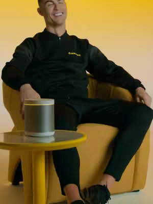 The cover image features a seated man, Cristiano Ronaldo, in a casual studio setting. He's wearing a black tracksuit and sitting in a yellow chair, with a side table holding a black and silver device. Ronaldo is smiling and looking off to the side. The background is a solid yellow gradient. Overall, the image has a modern and clean composition.