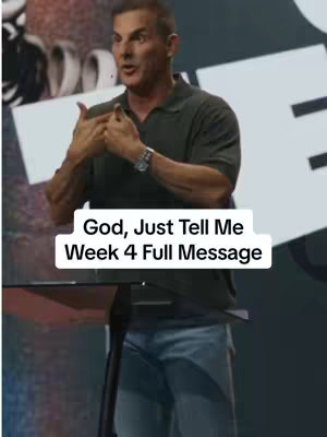 A man in a dark green shirt and blue jeans is standing behind a podium, gesturing with his hands. The background is blurred, with some blurred architectural elements visible. A white rectangle with black text overlays the bottom of the image.