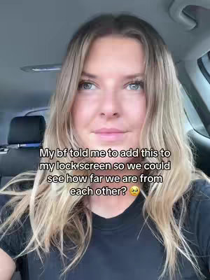 A selfie-style video shows a woman looking at the camera while inside a car. She has blonde hair and is wearing a black shirt. Text overlayed on the image indicates the woman's boyfriend told her to add something to her lock screen to track their distance.