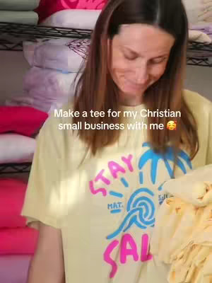 A woman stands smiling, holding fabric. She wears a light yellow t-shirt with a blue and pink graphic on the front. Behind her are shelves with stacked items.