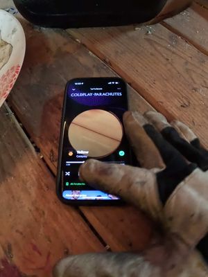 The image shows a close-up of a person's hand, covered in a work glove, holding a smartphone. The phone's screen displays the music app, Spotify, streaming ‘Yellow’ by Coldplay. The circular album art is visible, with a wooden surface in the background. A part of a blurred plate and what appears to be a black object is visible in the background.