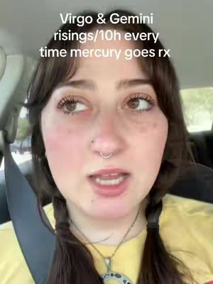 A selfie-style video with a woman looking slightly upwards and to the left, against a blurred background suggesting being inside a vehicle. The woman has dark hair in two braids and a nose ring. She is wearing a yellow shirt and a necklace. Text overlayed on the image reads, 'Virgo & Gemini risings/10h every time mercury goes rx'