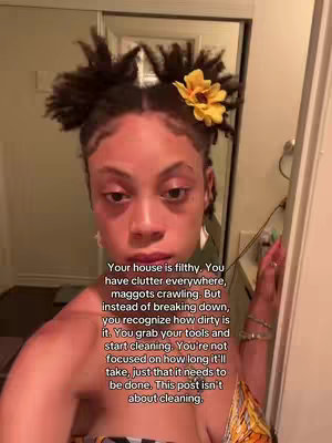 A woman with dark skin and curly hair is centered. She has two buns, with a yellow flower, and visible text overlays. She is facing the camera, with a bathroom door visible. The background is slightly blurred.