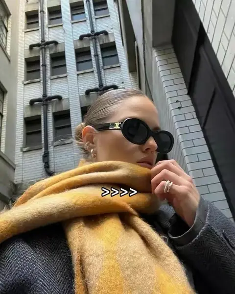 A woman wearing sunglasses and a yellow scarf poses outdoors. The background shows a tall building. The image has a warm color palette, featuring yellow, gold, and grey tones.