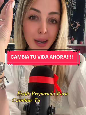 A woman with blonde hair and light skin is in the center of the frame, addressing the camera. A text overlay in a rectangular shape with a pink background reading 'CAMBIA TU VIDA AHORA!!!!' appears above the woman's head. Behind the woman is a slightly blurred background with a dark-colored backdrop, and some items in a shelving unit. A microphone with a red band and bow is in front, framing the woman’s face, below the hook text is the another overlay text 'Estás Preparado Para Cambiar Tu'.