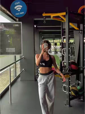A woman is taking a mirror selfie in a gym. She's in workout attire with headphones, holding a phone to take the picture. The background has gym equipment.