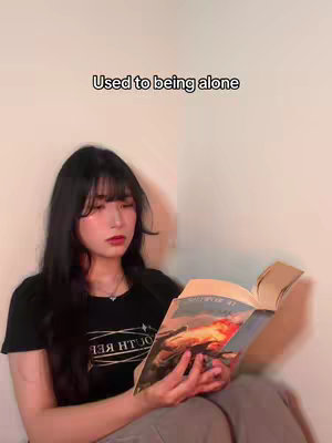 The image shows a young woman sitting and reading a book. She has long black hair with bangs, red lipstick, and is wearing a black t-shirt. The book is open, and the text overlay says "Used to being alone". The background is a neutral, light-colored wall.