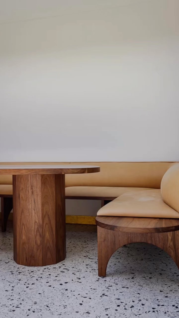 The image shows a modern and minimalist interior design, featuring a combination of a light-colored, curved banquette, a large, round wooden table, and a geometric-patterned floor. The banquette has a cream-colored cushioned seat and sleek wooden frame. Adjacent to the banquette is a matching wooden table with a round tabletop and a sturdy, cylindrical base. The flooring has a speckled pattern, adding texture to the scene. The background is a plain white wall, which enhances the clean and contemporary aesthetic