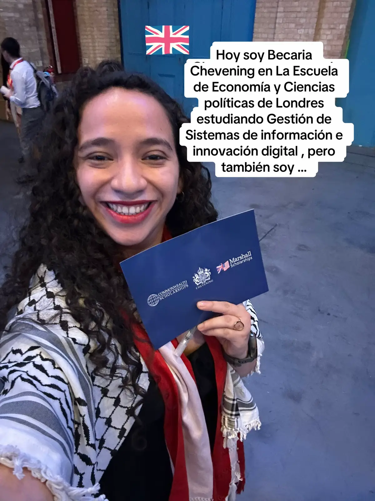 A selfie of a smiling woman holding a blue document and looking at the camera. Behind her is a blue door with a Union Jack flag sticker on it, suggestive of London, and text overlayed on the image.