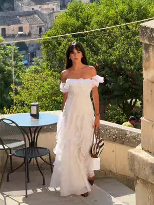 A woman in a white off-the-shoulder dress stands on a balcony with a cityscape backdrop. In the foreground, a round table and chair are visible. The woman holds a striped handbag. The scene is set on a sunny day.