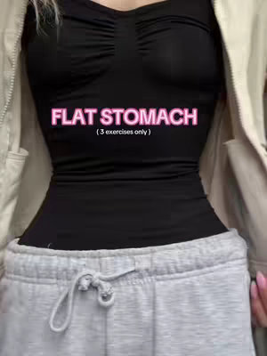 A woman is shown from the torso up, wearing a black top and gray sweatpants. The photo is cropped to focus on her midsection, where text overlays reading "FLAT STOMACH" in pink with "(3 exercises only)" beneath it. The woman's stomach appears flat, and her outfit is casual.