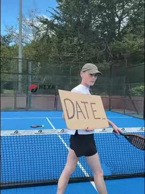 A young man stands on a blue court, holding a cardboard sign that reads "DATE." He wears a white t-shirt, black shorts, a cap, and sunglasses, with a padel court net and surrounding greenery visible in the background.