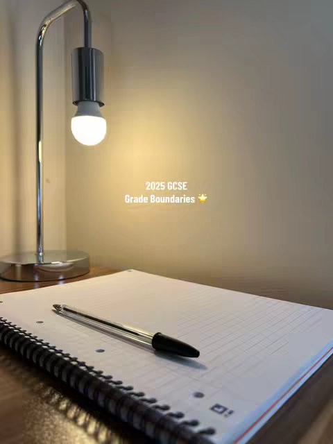 The image shows a notebook with a pen lying on it, lit by a desk lamp. The setting suggests a study environment. The background is a light-colored wall. The lamp has a chrome finish.