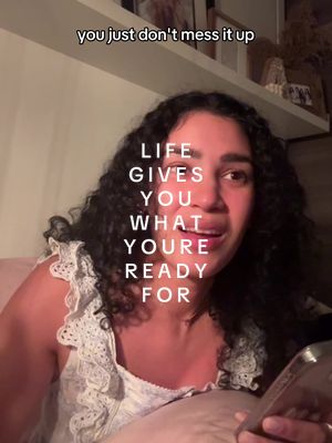 A woman with long, dark curly hair is looking at the viewer in a medium shot. She is wearing a white, lace-trimmed camisole. The background has a warm, inviting look with a soft glow. The text "LIFE GIVES YOU WHAT YOU'RE READY FOR" in a white, vertical font is superimposed on the image.