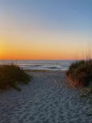 A serene beach scene at sunset. The sky transitions from a deep orange near the horizon to a light blue at the top. A sandy path leads towards the ocean, framed by dark green dune grass. The gentle waves reflect the sky's colors.