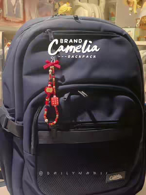 A close-up shot of a navy blue backpack. The backpack has a textured front pocket, a smaller top pocket, and a main compartment hidden by the angle. A charm composed of red bow, beads and various red, white, and yellow accents hangs from the zipper. Overlayed white text reads "BRAND Camelia --BACKPACK."