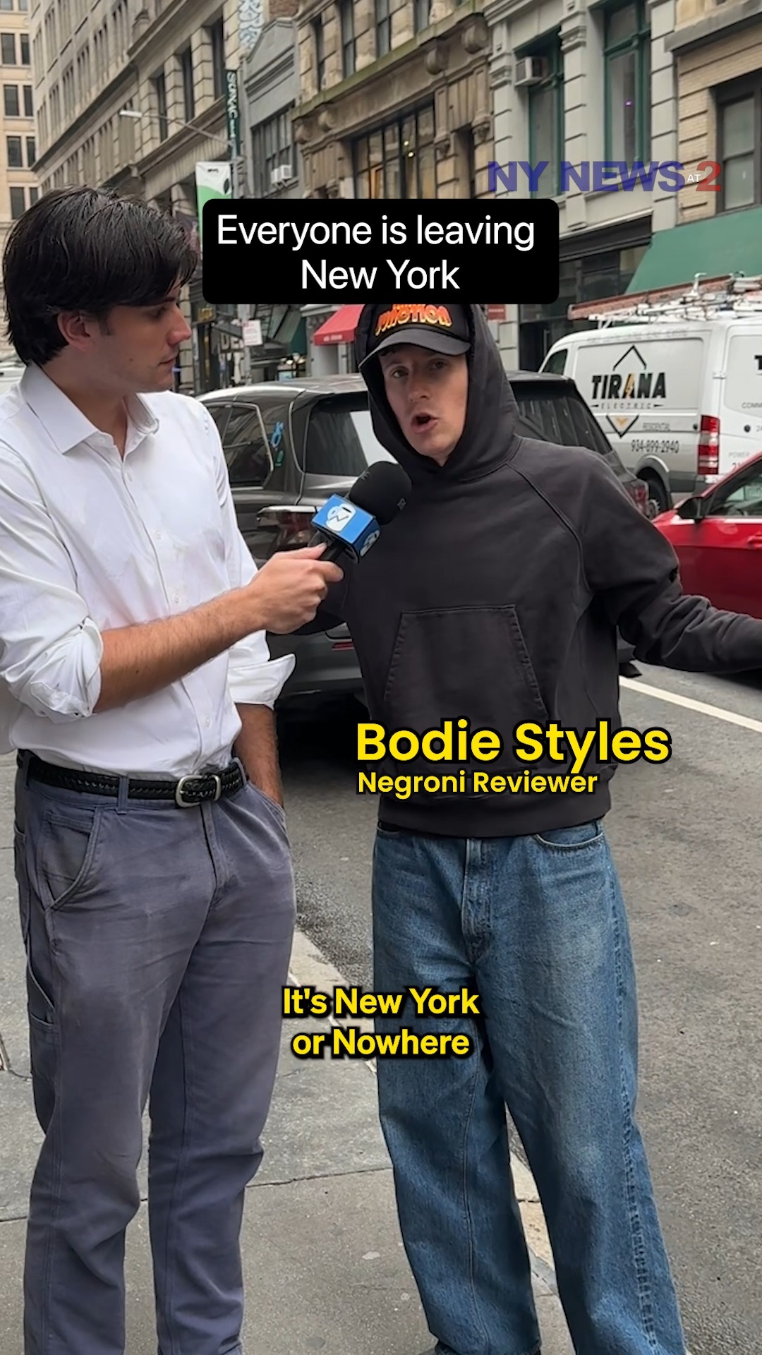 Two men are standing on a city street in front of building and parked cars. One man holds a microphone and is interviewing the other, who is the central focus of the image. Text overlays label them and provide context related to New York.