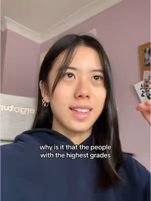 A young woman with long dark hair is the primary focus, captured from the chest up. She wears a dark blue hoodie and is positioned against a backdrop of a light purple wall and a white ceiling molding. Text overlayed at the bottom reads, 'why is it that the people with the highest grades'.