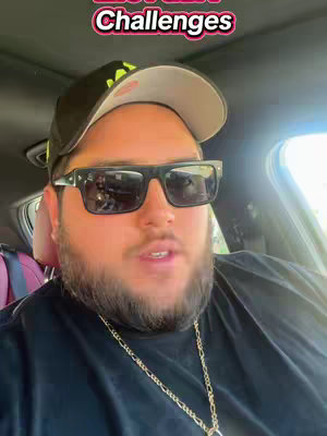 A man wearing a black baseball cap, sunglasses, and a gold chain sits in a car. The car's interior is visible, with a purple seat.