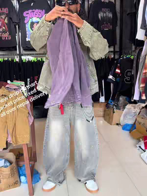 A man holding up and inspecting a purple jacket in a clothing store. The man wears sunglasses, a white cap, and a green jacket over a black t-shirt. He is standing in front of racks of clothing.