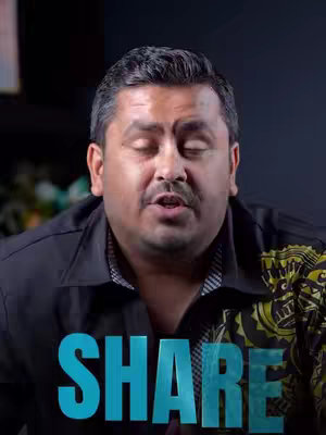 A medium shot features a man with short, dark hair slightly receding. He's wearing a black button-down shirt over a white collared shirt, and a patterned design is visible on his right shoulder. He is centered in the frame and speaking with his eyes closed. Turquoise text overlays the bottom, reading SHAR with the last E being cut-off.