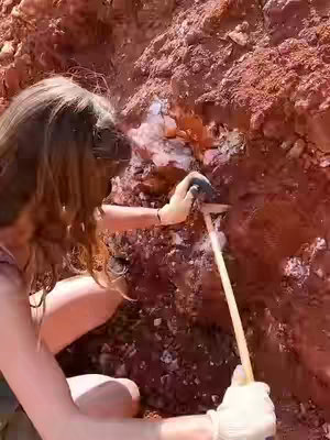 A woman is shown crouched next to a rock face, wearing gloves and using a tool to work on the rock. The setting appears to be an outdoor environment with reddish-brown soil and rocks.
