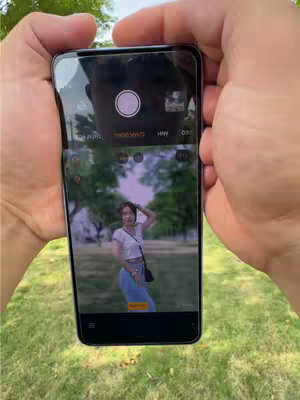 A person holding a smartphone displaying a woman posing for a portrait in a park. The phone screen shows the woman styled in casual attire in front of a tree-lined path, with a blurred background. The person holding the phone, whose hands are visible, is in focus. The phone's camera interface, with various control icons across the edges, fills the rest of the frame. The outdoor scene and warm lighting colors are visible, suggesting a sunny day. 