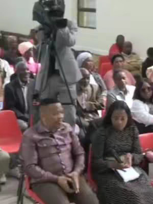The cover image shows a scene inside a building, possibly a church or event hall, with a group of people seated. A camera operator is positioned in the foreground, recording the scene. In the front row, two people are prominently featured, a man wearing a purple shirt with a shaved head and another woman in a black dress with a microphone and a notepad. Other seated people are in background, possibly the audience. The lighting appears to be natural or overhead, and the overall composition suggests the recording of a speech or testimony.