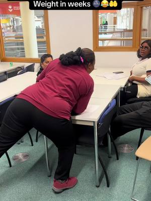 A woman in a maroon shirt and black pants leans against a table, with two other women in the background. The room is bright with large windows.