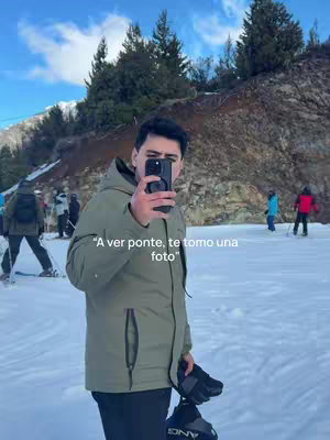 A man in a green jacket takes a selfie with a phone held in front of his face. He is standing in a snowy area with trees and other people in the background. Text on the image says, "A ver ponte, te tomo una foto".