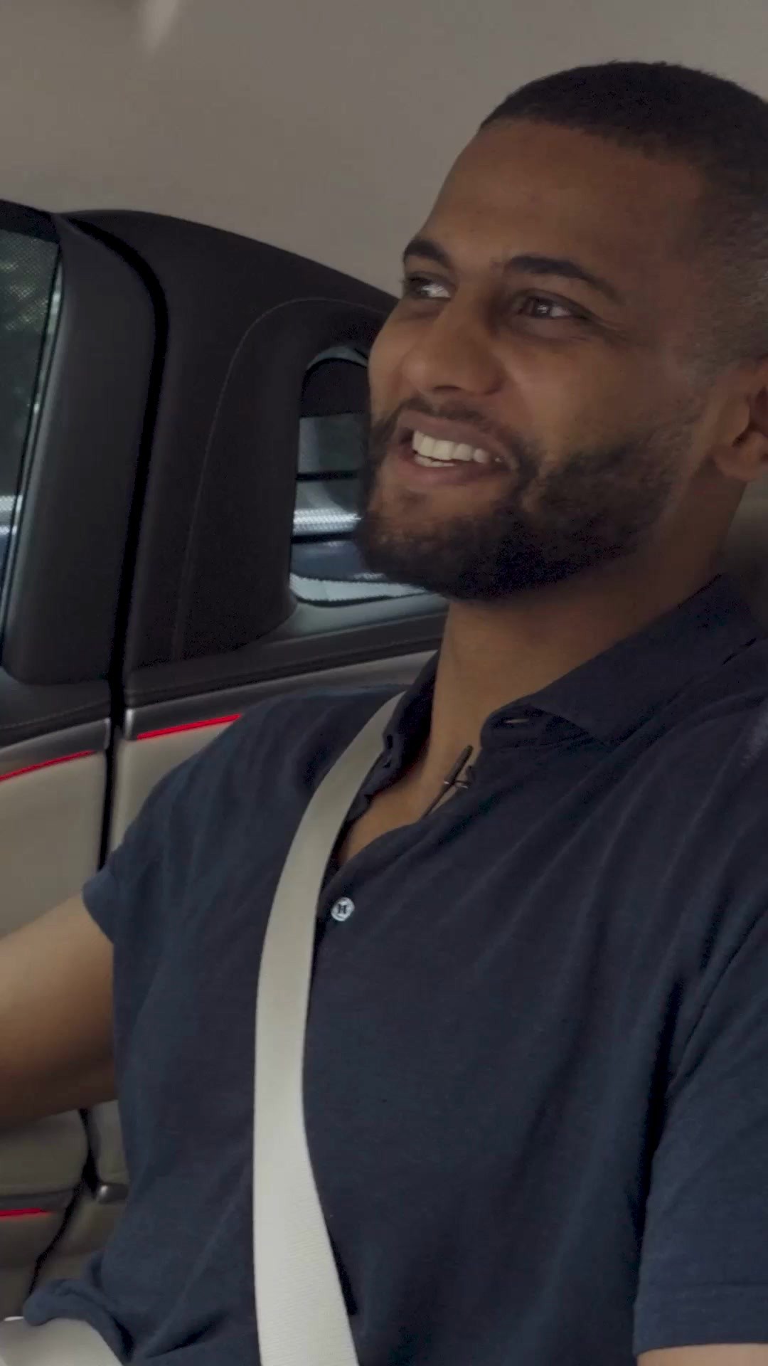 The cover image shows a man in the passenger seat of a car, smiling broadly with his head turned toward the camera. He is wearing a navy blue polo shirt and a seatbelt. The vehicle interior is visible, with a dark headliner overhead and glimpses of the car's interior details including a red accent light. The lighting appears to be natural.