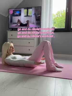 A woman is lying on her back on a pink exercise mat wearing a white long-sleeve shirt and pink pants. She's in what appears to be a home setting, with a white dresser and a large-screen TV showing a workout video behind her. The hook text is overlaid on the image: 'go and do nikola's workout'.