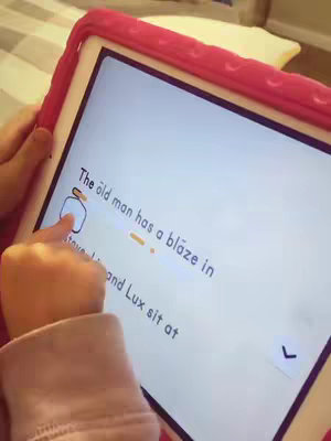 A close-up shot of a hand holding a tablet showing an educational app. The screen displays a sentence with highlighted words, likely for reading practice. The tablet is encased in a pink protective cover, suggesting a child-friendly device. The soft focus and warm colors create an inviting atmosphere, with the focus on the learning activity.