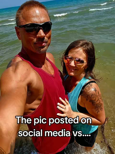 A couple stands in the water at the beach, posing for a photo. The man, on the left, is wearing a red tank top and sunglasses. The woman is on the right, with a blue top, also wearing sunglasses. Text overlayed across the bottom reads, 'The pic posted on social media vs....'