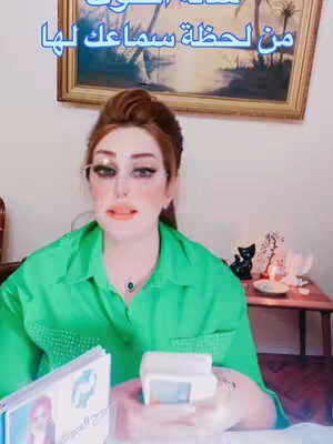 A woman with red hair wearing a green shirt is seated in front of a camera, holding a small white device (possibly a card reader). She has makeup on, including prominent eye shadow. Behind her is a framed print, and on a small table to the right, there's a black cat figurine and a candle.