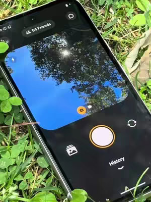 A close-up shot of a smartphone displaying the Locket app interface, set against a backdrop of vibrant green grass with some clovers. The screen shows a view of the sky through some tree branches, and the app interface is visible with buttons and icons. The phone is positioned at a slight angle, offering a clear view of the screen's design.