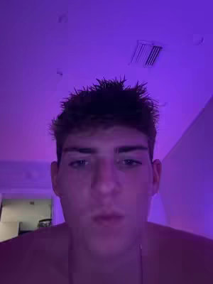 A close-up shot of a young man's face, illuminated by a purple light. The background is slightly blurred and shows the ceiling of a room. The man is looking directly at the camera.